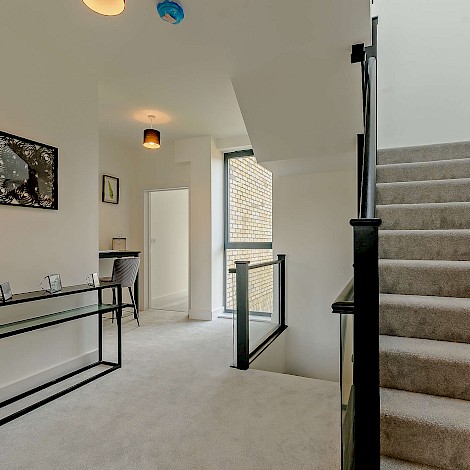 Interior hall way view of a Connaught Barracks property showing grey carpeted staircase with black banister and hallway with parquet light cream carpet and modern black furniture and accessories